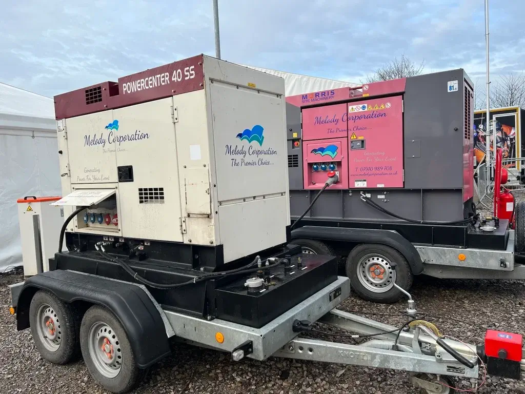 Two portable generator units on trailers, one white and one purple, parked on gravel next to a white tent under a cloudy sky.